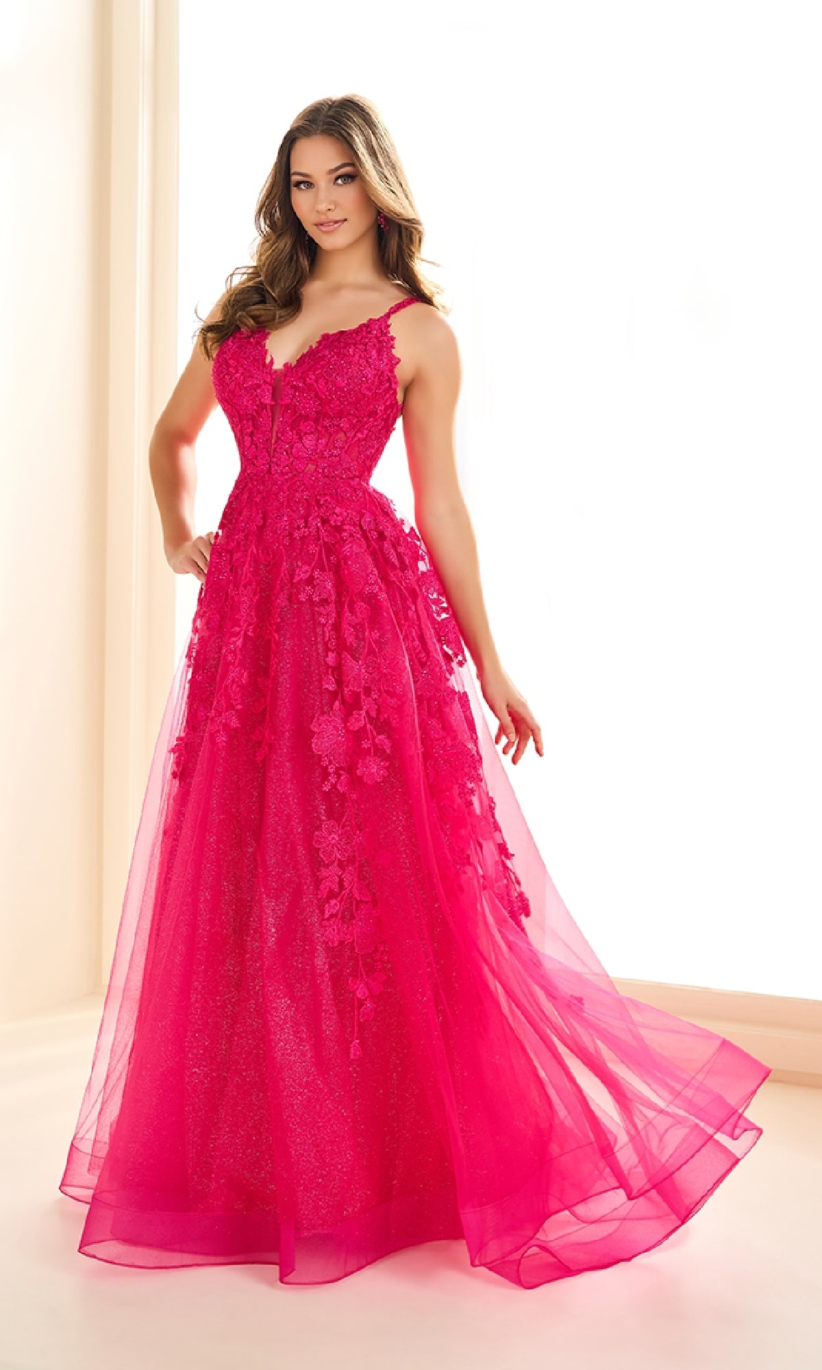 A woman stands in a bright room wearing the Ellie Wilde EW36015, a sleeveless, floor-length hot pink prom dress with a corset bodice, floral appliques, and a sheer overlay.